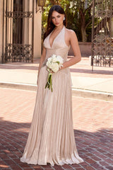 A woman stands outdoors on a brick walkway wearing a sleeveless, halter-neck, floor-length champagne pleated gown and holding a bouquet of white flowers.