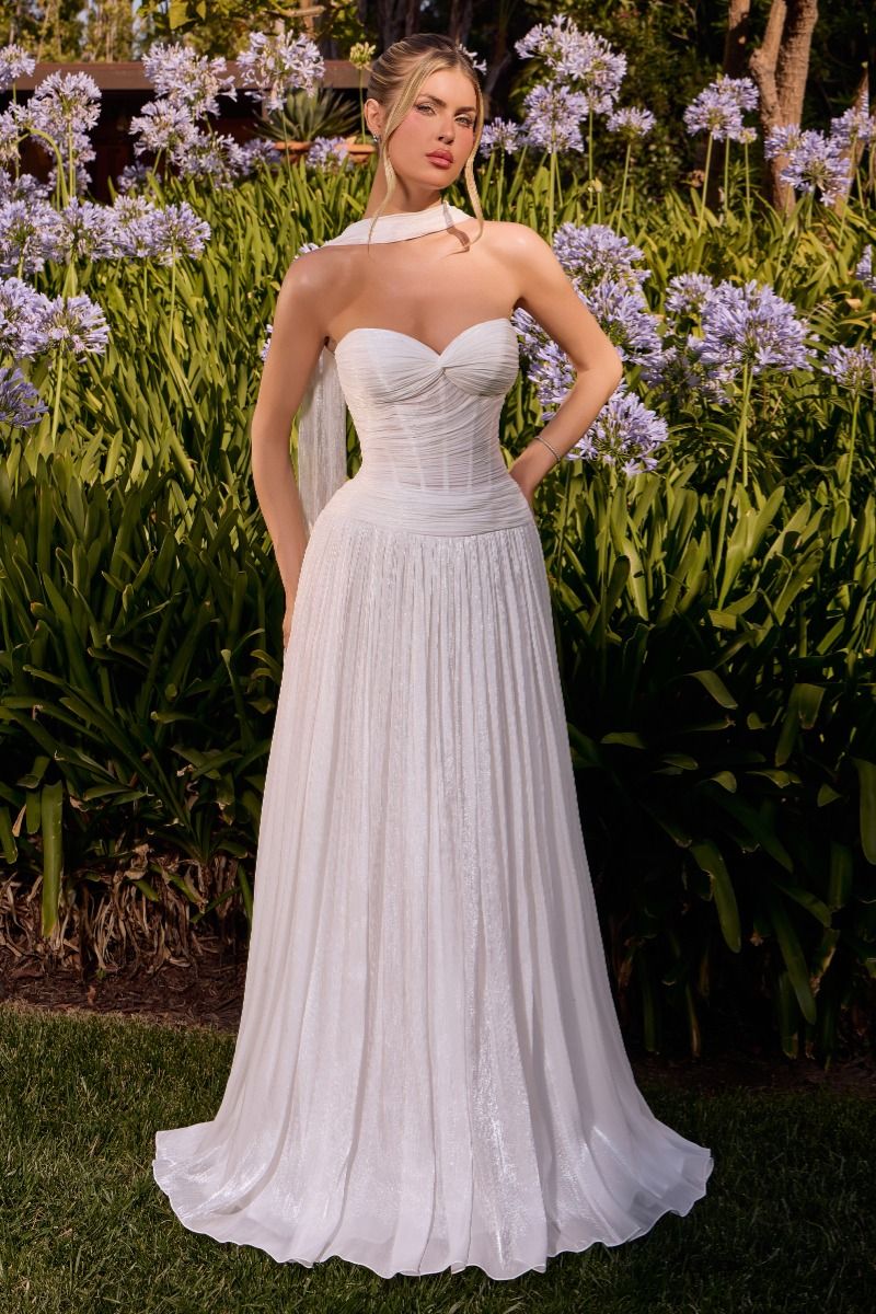 A woman wearing a strapless white wedding gown stands outdoors in a garden with green foliage and purple flowers behind her. The dress has a fitted sweetheart bodice, a sheer illusion neckline, and a full flowing skirt.