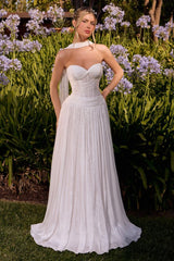 A woman wearing a strapless white wedding gown stands outdoors in a garden with green foliage and purple flowers behind her. The dress has a fitted sweetheart bodice, a sheer illusion neckline, and a full flowing skirt.