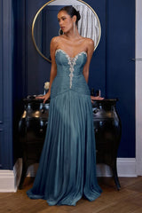 A woman models a floor-length strapless gown in a dusty teal blue, featuring a sweetheart neckline, a beaded embellished bodice, and a pleated skirt. She is posed in front of a dark wood console table and a blue paneled wall with a gold-framed mirror.