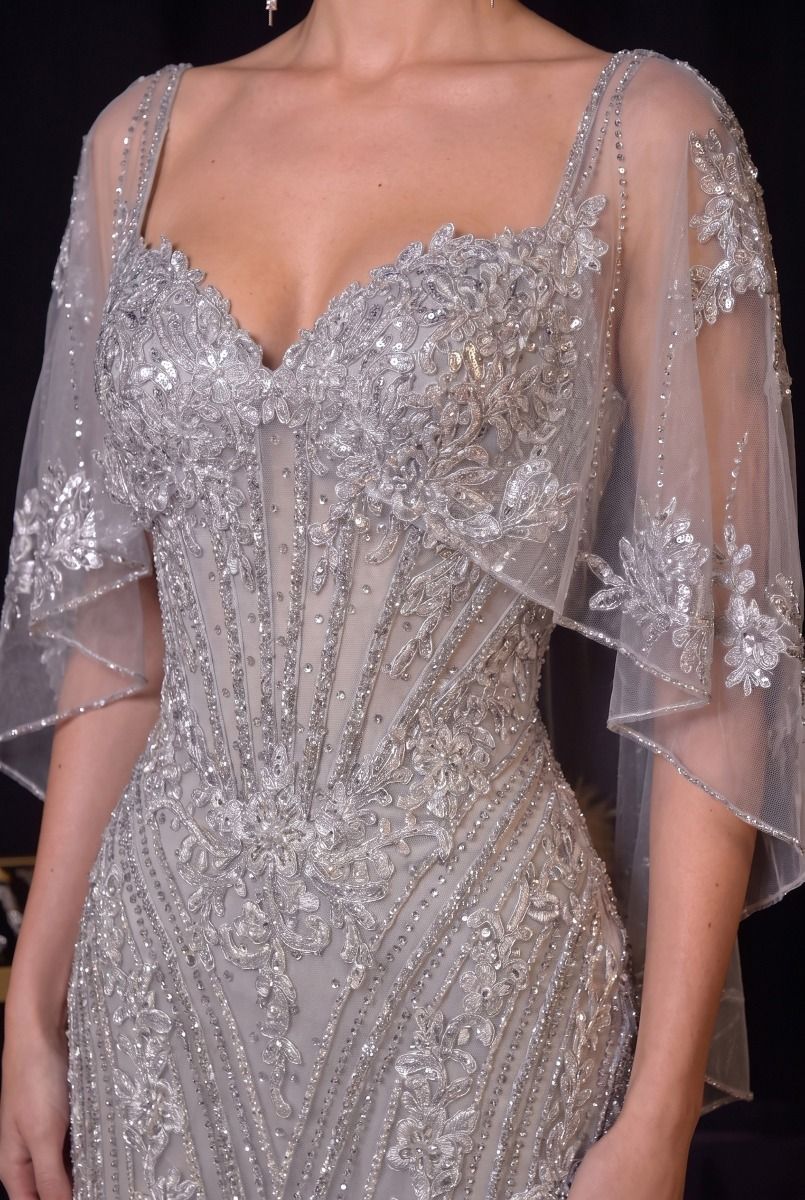 A close-up of a model wearing a heavily embellished silver gown with intricate beadwork, sequins, and sheer flutter sleeves against a dark background.