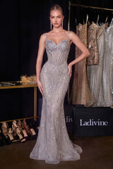 A model wears a floor-length, silver embellished evening gown with spaghetti straps and a sweetheart neckline in a runway-style showroom.