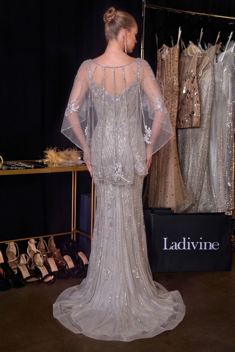 A model from the back wearing a heavily embellished silver lace and sequin evening gown with sheer draped cape sleeves, standing in a bridal boutique. 