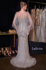 A model from the back wearing a heavily embellished silver lace and sequin evening gown with sheer draped cape sleeves, standing in a bridal boutique. 