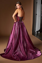 A woman modeling a strapless ball gown from the back, featuring a deep V-neckline, sheer lace corset bodice, and a voluminous satin skirt in a rich plum purple color, set against a warm brown studio backdrop.