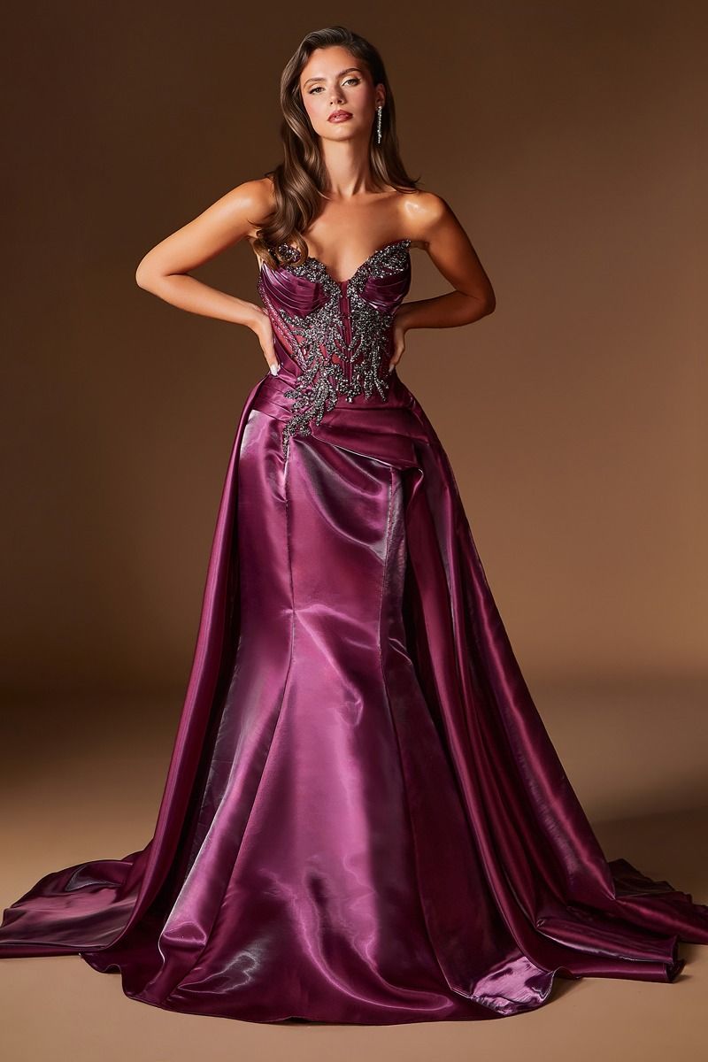 A woman modeling a floor-length formal gown in rich plum purple satin with a heavily embellished beaded bodice and a full skirt, posed against a warm brown studio backdrop.