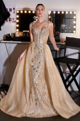A woman modeling a floor-length champagne beige ball gown with intricate silver and gold beaded embellishments, standing in a dressing room with a mirror and studio lights in the background.