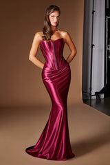 A woman modeling a strapless mermaid gown made of shiny satin-like fabric in a rich magenta-pink color, set against a warm brown studio backdrop.