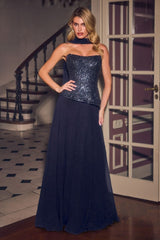 A woman modeling a strapless navy blue evening gown with a sequined corset-style bodice and a flowing tulle skirt, standing indoors near a staircase and glass-paneled door.