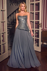 A woman models a strapless floor-length gown with a heavily embellished silver sequined bodice and a flowing slate gray skirt, standing in a doorway of an elegant interior.