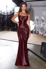 A woman models a floor-length, satin-like burgundy evening gown with a draped bodice and square neckline in a studio setting with lighting equipment and a chandelier visible in the background.