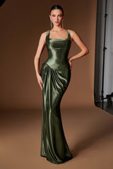 A woman models a floor-length olive green satin gown with a square neckline, structured bodice, and draped skirt against a warm brown studio backdrop.