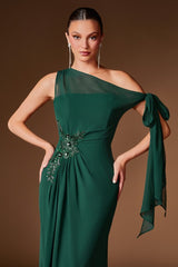A woman modeling a dark green one-shoulder evening gown with sheer mesh fabric, a side tie, and embellished floral embroidery at the waist, set against a solid brown studio background.