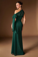 A woman modeling a floor-length emerald green one-shoulder gown with a draped bow detail on the bodice, photographed against a warm brown studio backdrop.