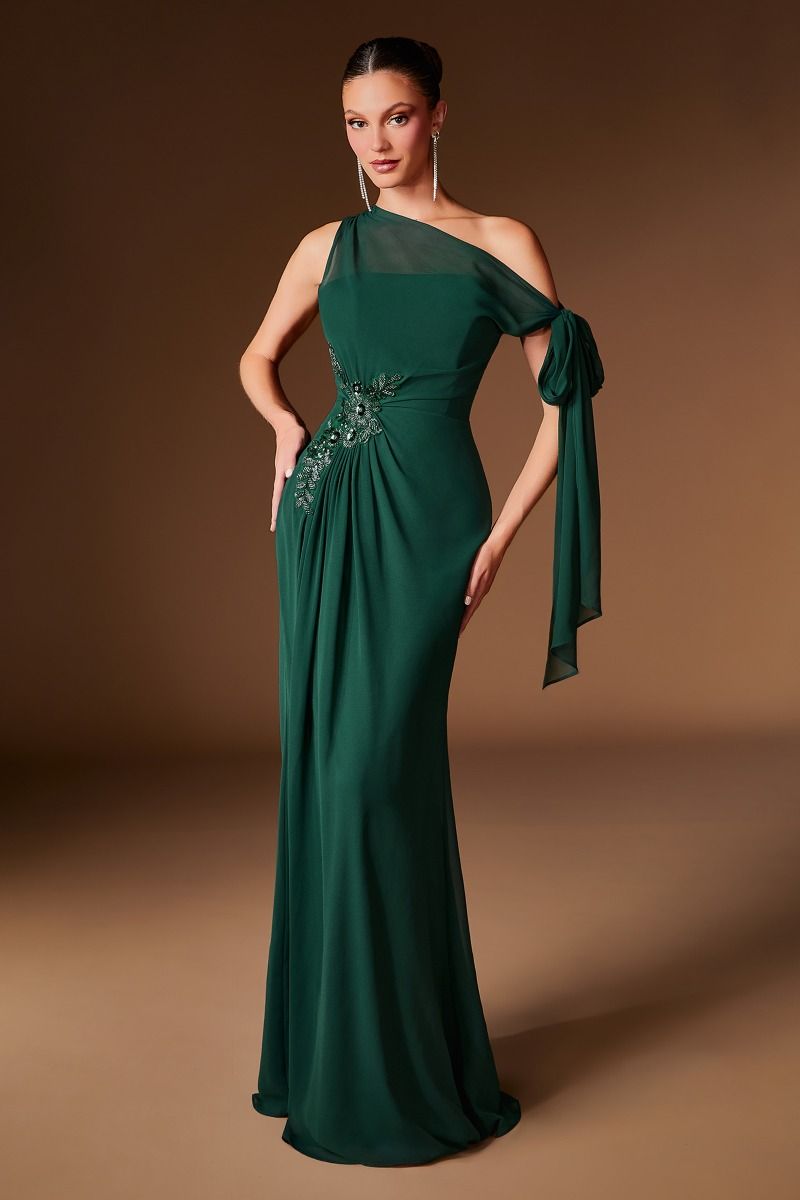A woman modeling a floor-length emerald green one-shoulder evening gown with a sheer mesh shoulder panel, draped tie detail, and embroidered embellishment at the waist, set against a warm brown studio backdrop.