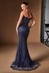 A woman modeling a strapless navy blue evening gown with shimmering vertical beadwork and a sheer tulle overskirt, photographed in a studio with a warm brown backdrop.