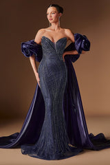 A woman models a strapless evening gown in deep navy blue with textured vertical beadwork and dramatic puffed sleeves, posed against a warm brown studio backdrop.