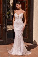 A woman modeling a strapless white mermaid-style gown with a deep V neckline, fitted bodice, and flowing skirt, standing in front of dark wooden doors and a beige stucco wall.