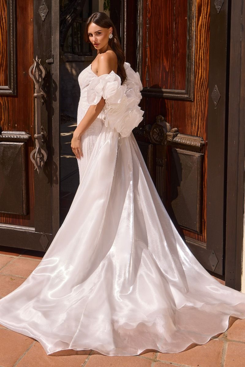 A woman wearing a white off-the-shoulder wedding gown with dramatic ruffled sleeves and a full satin train, standing in front of dark wooden doors.