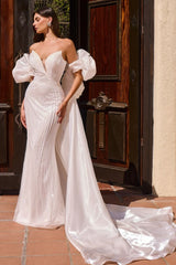 A woman wearing a strapless white wedding gown with dramatic puff sleeves, a fitted pleated bodice, and a long satin train, standing outdoors in front of a dark wooden door.