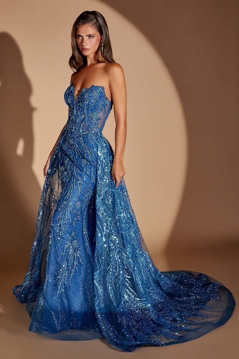 A woman modeling a strapless royal blue ball gown with intricate sequin and bead embellishments, standing against a warm beige studio backdrop with dramatic spotlighting.
