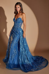 A woman modeling a strapless royal blue ball gown with intricate sequin and bead embellishments, standing against a warm beige studio backdrop with dramatic spotlighting.