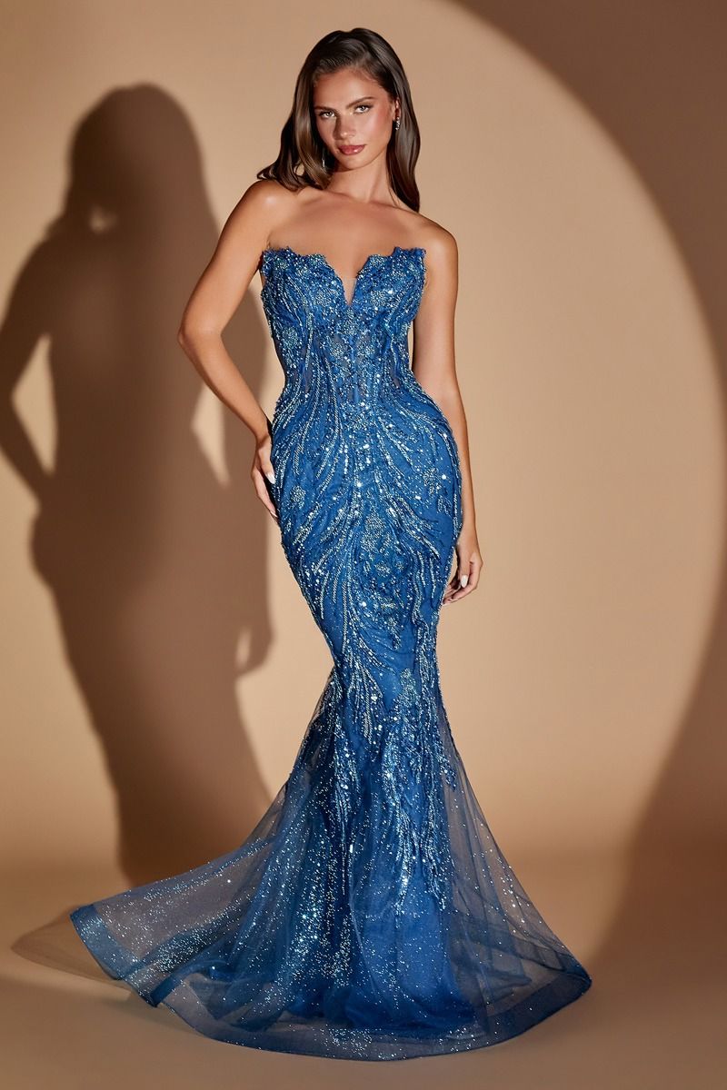 A woman models a strapless royal blue mermaid gown covered in shimmering sequins and beadwork, set against a warm beige studio backdrop with a soft spotlight and shadow.