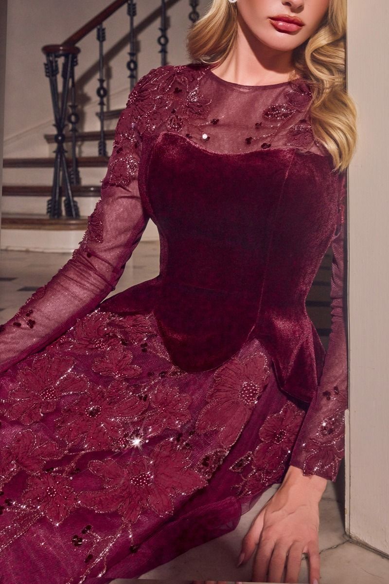 A woman wearing a long-sleeve burgundy velvet and mesh dress with floral lace appliques and sequins, posed indoors near a staircase.