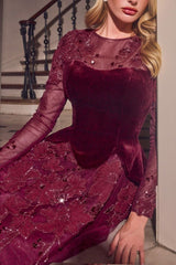 A woman wearing a long-sleeve burgundy velvet and mesh dress with floral lace appliques and sequins, posed indoors near a staircase.