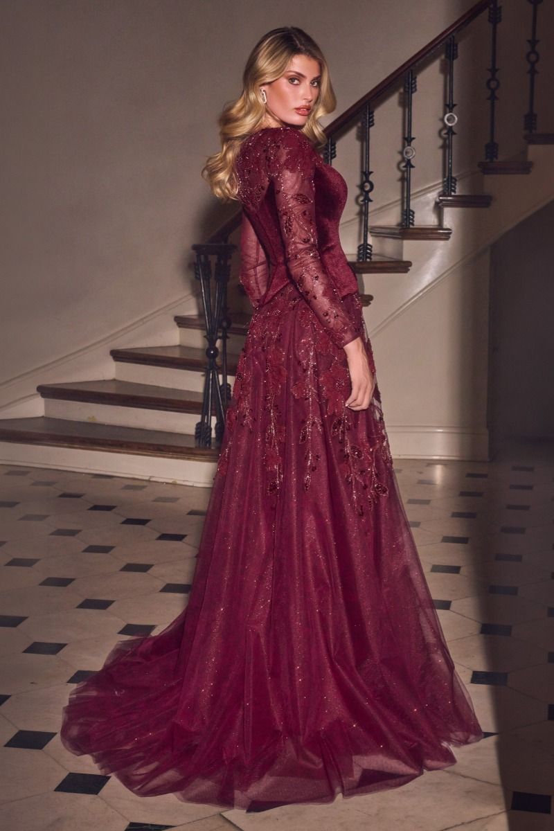 A woman wearing a long-sleeved burgundy tulle evening gown with lace and sequin embellishments, standing in a grand staircase hallway.