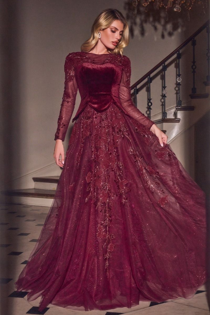 A woman models a floor-length burgundy ball gown with long sheer sleeves, a velvet bodice, and a voluminous tulle skirt with floral embroidery, set against an elegant indoor staircase background.