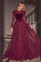 A woman models a floor-length burgundy ball gown with long sheer sleeves, a velvet bodice, and a voluminous tulle skirt with floral embroidery, set against an elegant indoor staircase background.