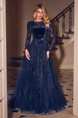 A woman models a floor-length navy blue ball gown with long sheer sleeves, a velvet bodice, and a voluminous tulle skirt with floral lace applique, standing in a doorway with a staircase in the background.