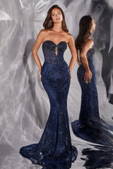 A woman models a strapless navy blue mermaid gown with a sweetheart neckline, sheer lace bodice, and a fitted beaded skirt. A mirrored view of the model and dress appears in the background against a draped gray fabric backdrop.
