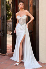 A woman models a strapless white evening gown with a sweetheart neckline, heavily embellished beaded corset bodice, high side slit, and a long flowing train, posed in front of a rustic wooden doorway.