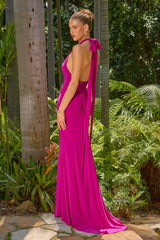 A woman wearing a bright fuchsia pink halter-neck floor-length gown with a draped back and long train, standing outdoors in a tropical garden with palm trees and lush greenery.