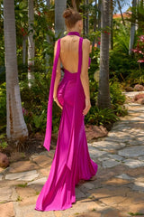 A woman wearing a bright magenta formal gown with a backless open back and long draped side panels, photographed outdoors on a stone path surrounded by palm trees and tropical greenery.