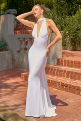 A woman modeling a floor-length white evening gown in an outdoor brick courtyard with stone steps and greenery in the background.