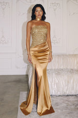 Woman wearing a gold strapless gown with a sequin-embellished corset bodice, draped satin skirt, thigh-high slit, and lace-up back.
