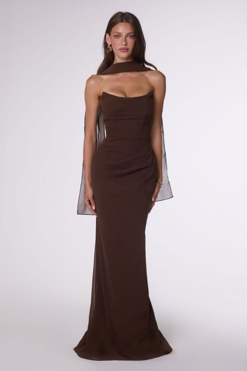 A woman modeling a dark brown strapless gown with sheer draped cape sleeves against a light gray studio background.
