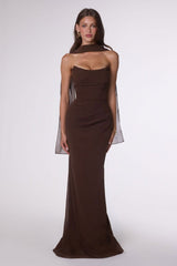 A woman modeling a dark brown strapless gown with sheer draped cape sleeves against a light gray studio background.