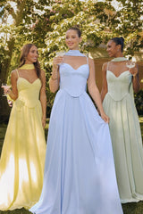 Three women standing outdoors wearing pastel floor-length corset gowns — one in light yellow with a flowing scarf detail, one in baby blue with a halter neck strap, and one in sage green with thin straps — posing in a garden setting