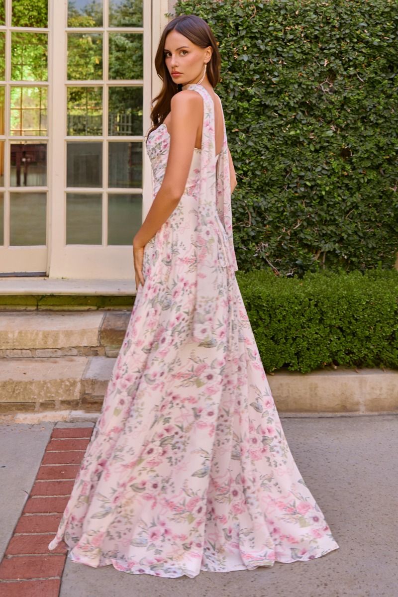 A woman wearing a sleeveless, floor-length floral gown with a tiered back, standing outdoors in front of a green hedge and white-framed glass door.