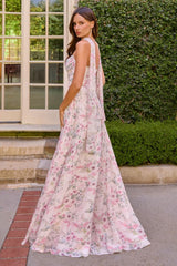 A woman wearing a sleeveless, floor-length floral gown with a tiered back, standing outdoors in front of a green hedge and white-framed glass door.