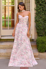 A woman wearing a strapless white maxi dress with a pink and green floral print, standing on stone steps in front of a building with shutters and greenery.
