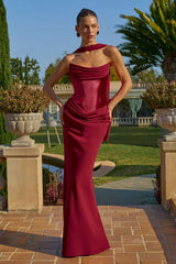 Woman in a burgundy strapless gown standing outdoors with greenery and a building in the background