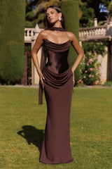 Woman in a brown evening gown standing outdoors on grass with trees and flowers in the background