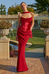 A woman stands outdoors on a brick patio in front of a large house and lawn, wearing a fitted, floor-length red gown with a draped halter-style neckline and ruched detailing.