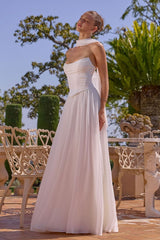 Woman in a white strapless gown standing outdoors with greenery and a clear sky.