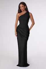 A woman modeling a floor-length black one-shoulder gown with a draped, gathered skirt against a light gray studio background.